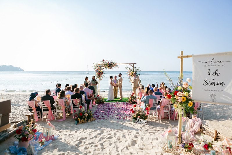 A beach wedding ceremony on Phuket with a floral arch and seated guests at the shoreline.